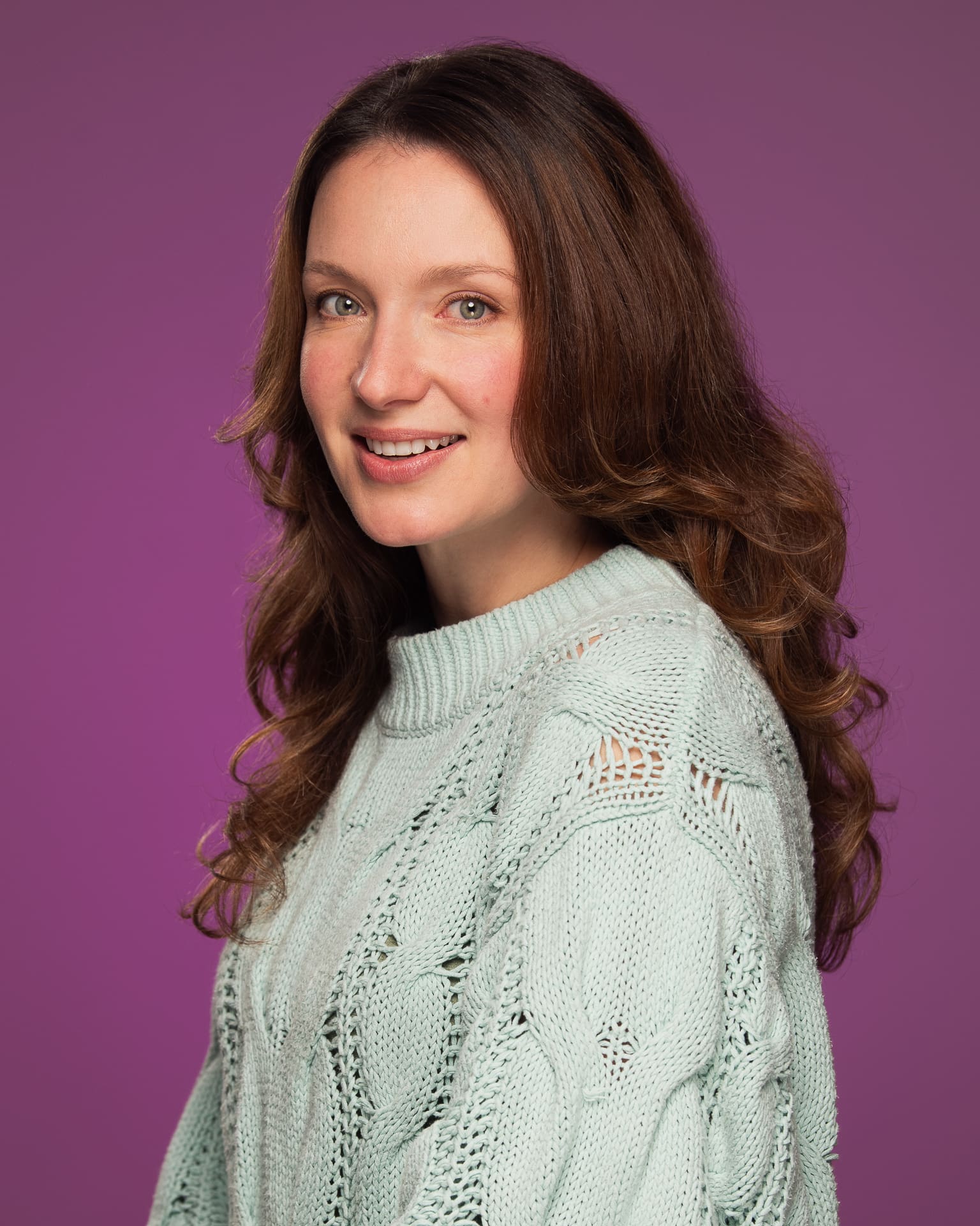 Woman smiles softly to camera on a purple/plum coloured background.