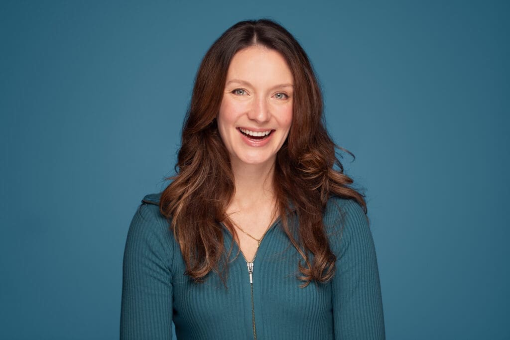 Woman smiles widely to camera. She wears teal and is photographed on a similar background.