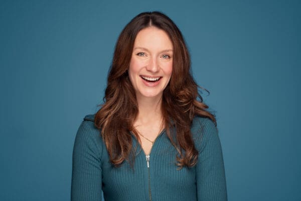 Woman smiles widely to camera. She wears teal and is photographed on a similar background.