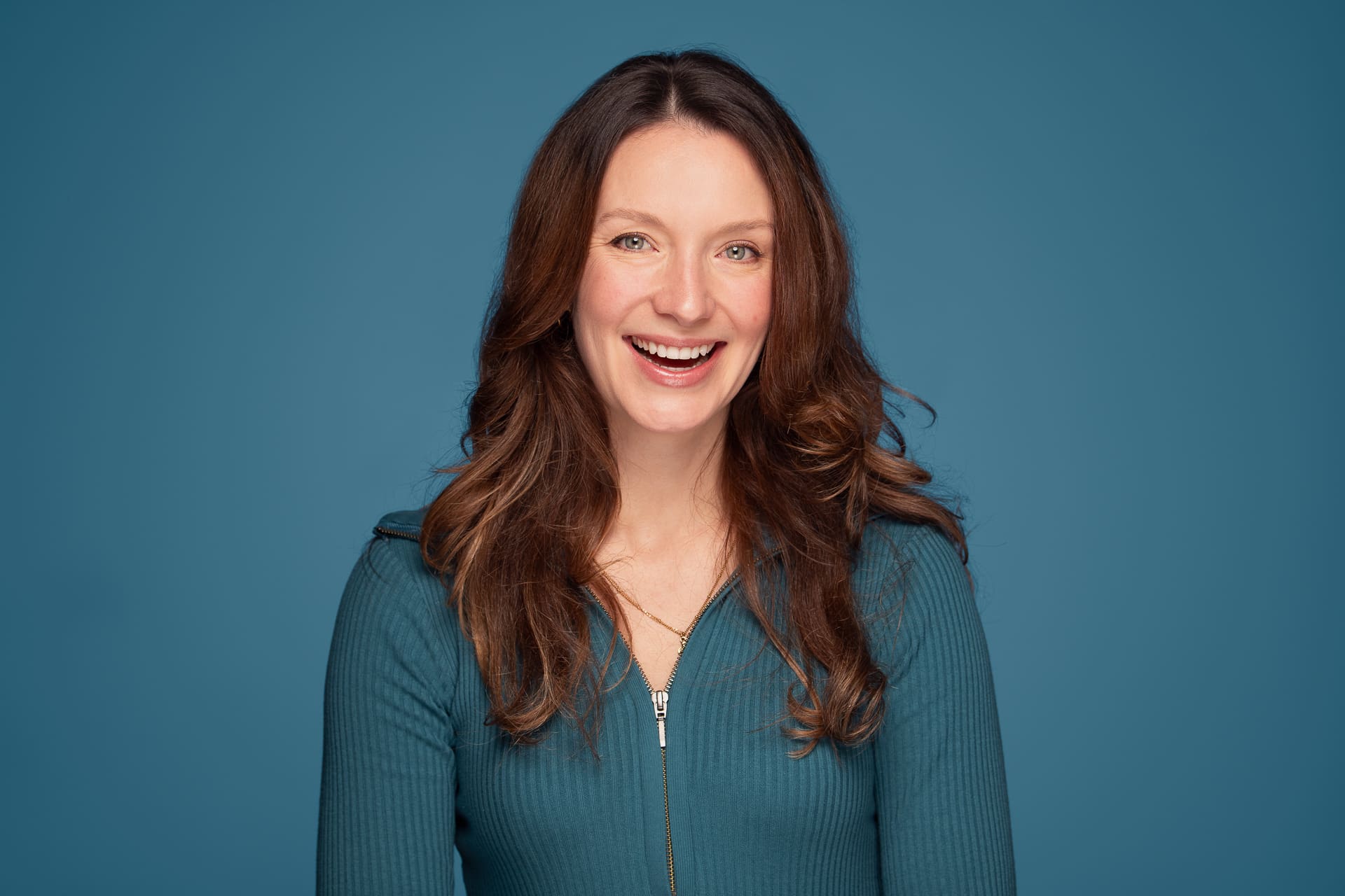 Woman smiles widely to camera. She wears teal and is photographed on a similar background.