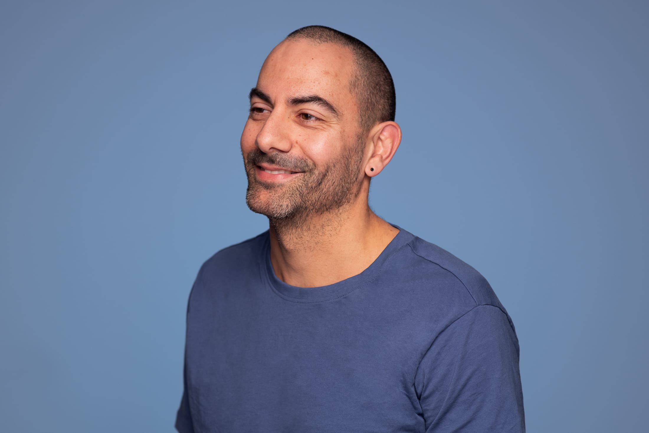 Man smiles away from the camera, wearing a blue tshirt on a blue background.