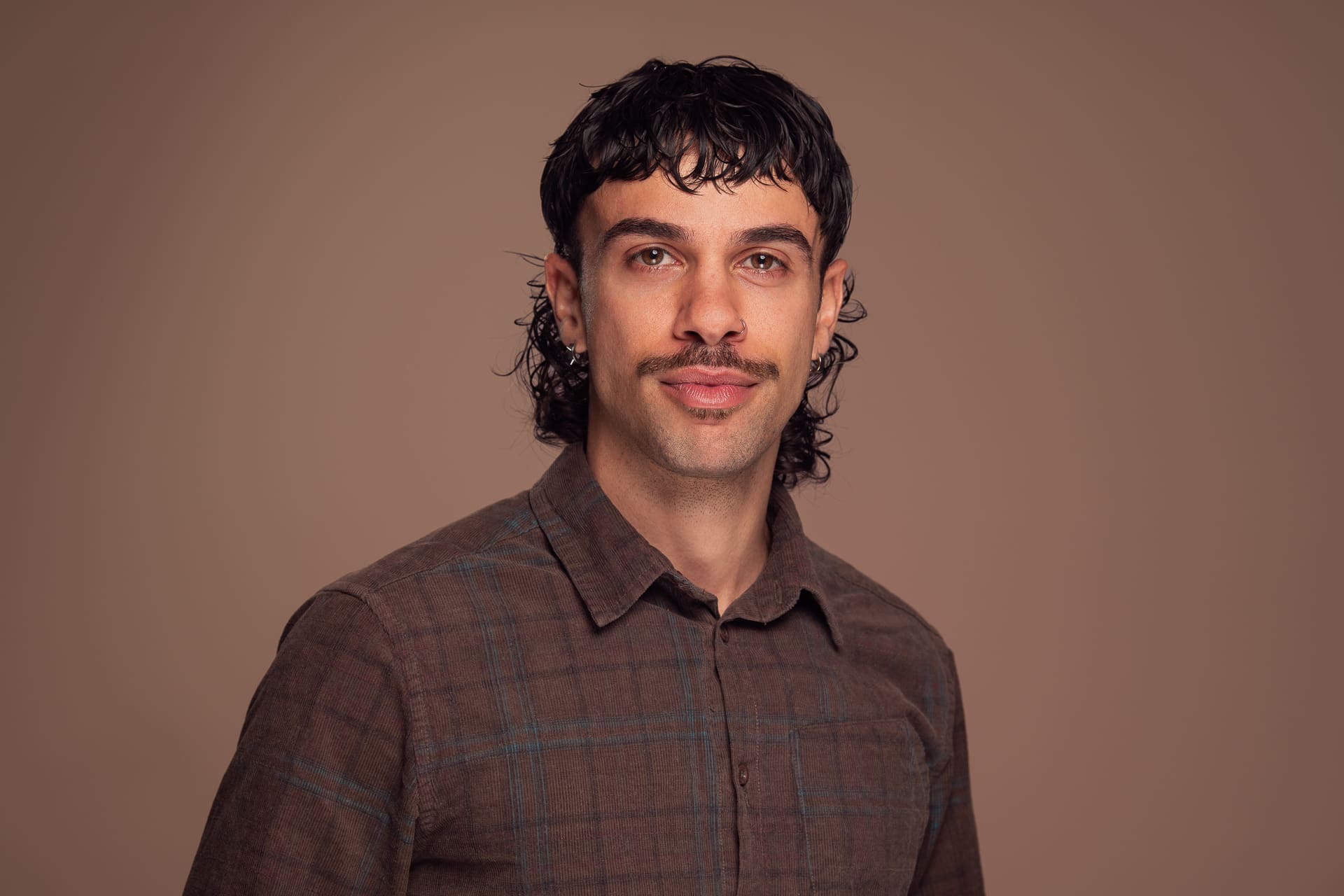 Man looks to camera with a soft expression. he wears a brown button up shirt and is photographed on a latte brown background.