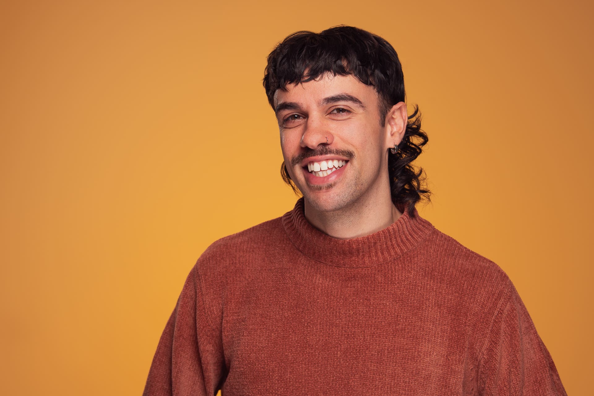 Man smiles to camera in a burnt orange jumper. The background is a dark yellow.