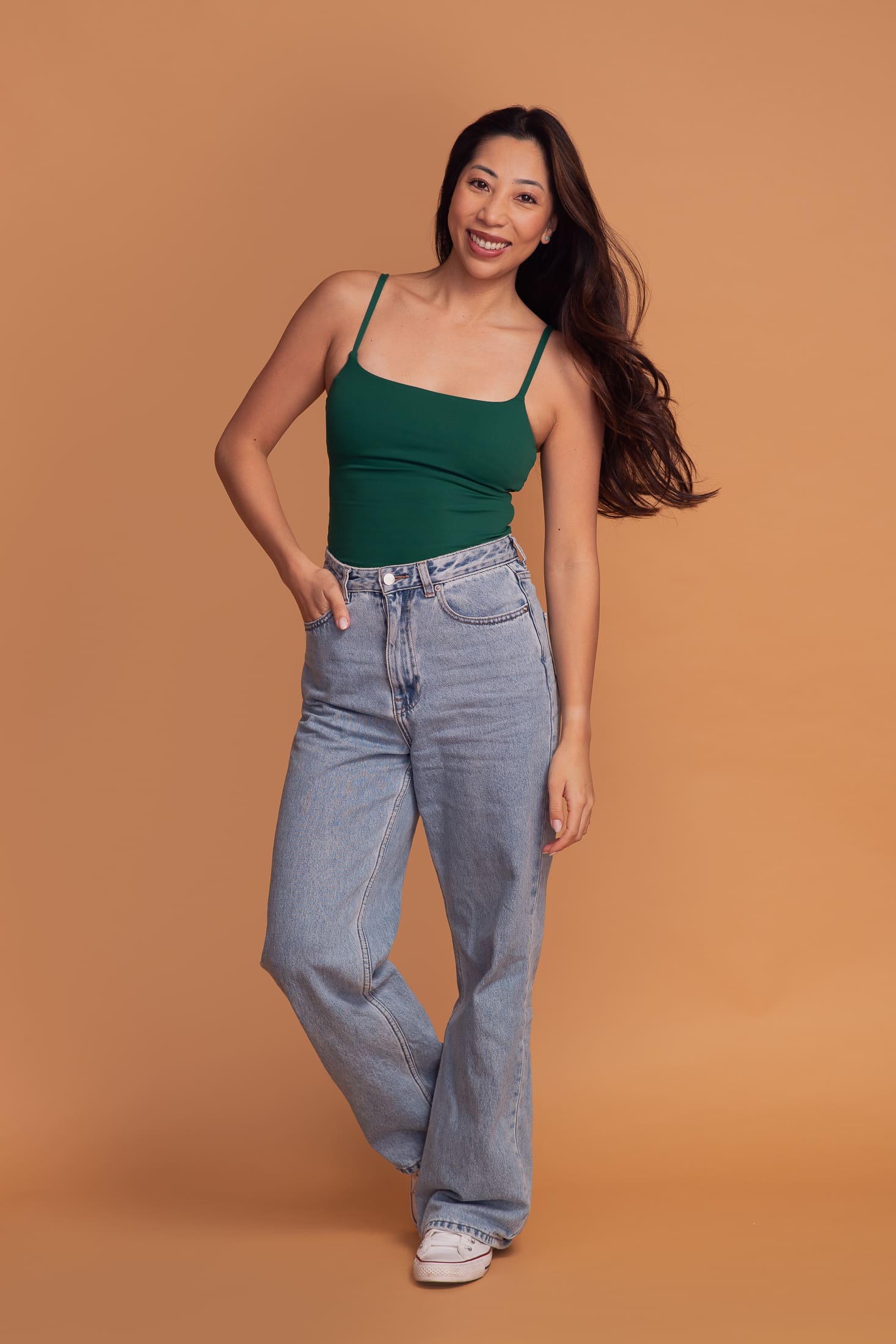 Full length portrait of a woman smiling and standing on a soft orange background. She has one hand in the pocket of her jeans.