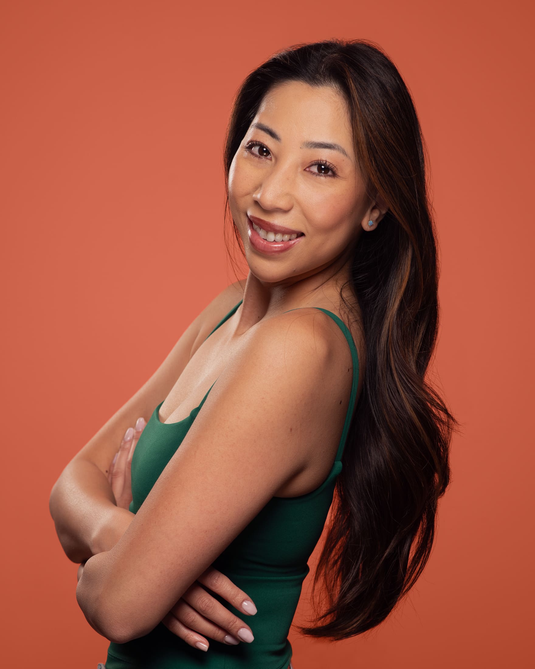 Woman looks to camera with arms crossed. She is positioned side on, and you can see her long hair. The background is a deep orange.
