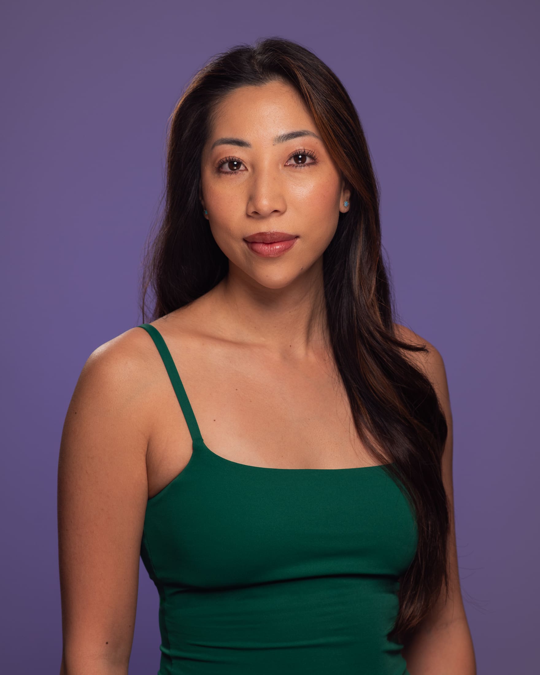 Woman loks to camera with soft, neutral expression. The background is purple and her top is green.