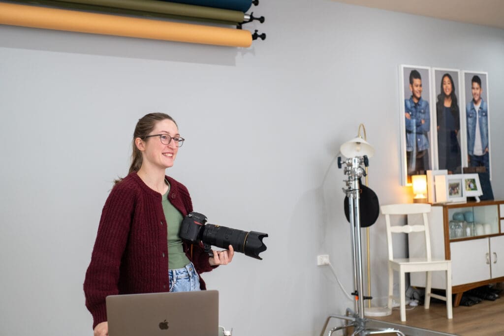 Julia holds her camera and stands next to her laptop in her studio, smiling away from the camera.
