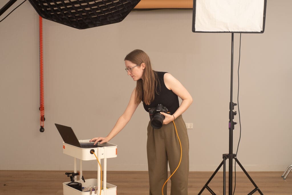 Julia interacts with her laptop and holds her camera in behind the scenes image.